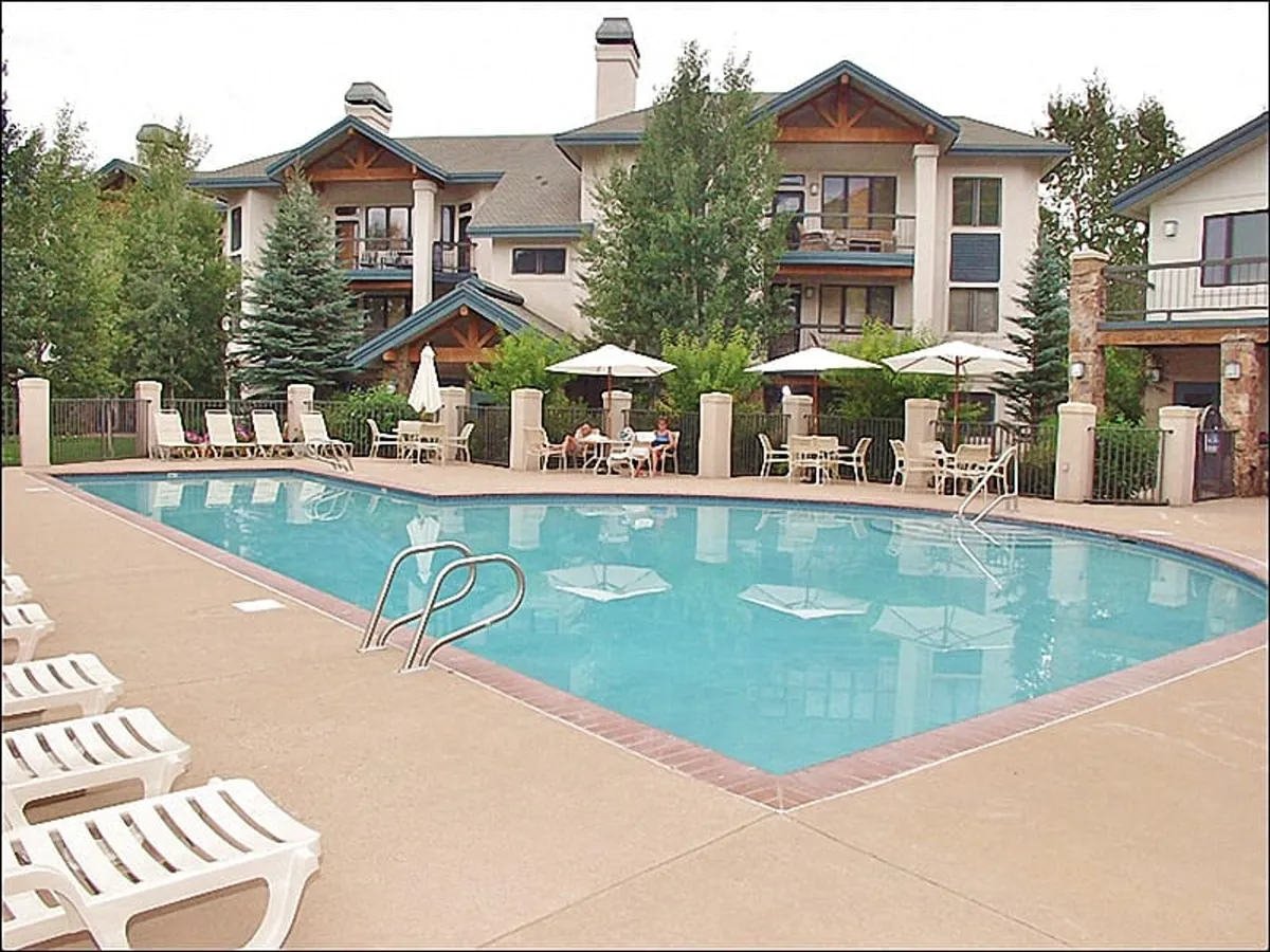 A sunlit outdoor swimming pool surrounded by lounge chairs, umbrellas, and lush greenery, with the building's architecture visible in the background. The atmosphere is relaxed and inviting, perfect for swimming, sunbathing, or gathering with friends.