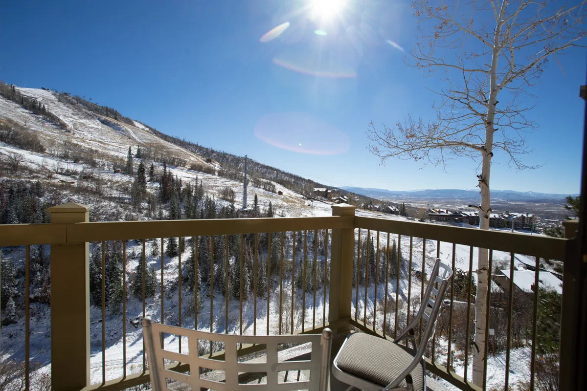A serene exterior view from a balcony overlooking snow-covered mountains, with sunlight glinting off the slopes. The space invites relaxation and enjoyment of the natural beauty, suggesting a perfect spot for unwinding or watching skiers.