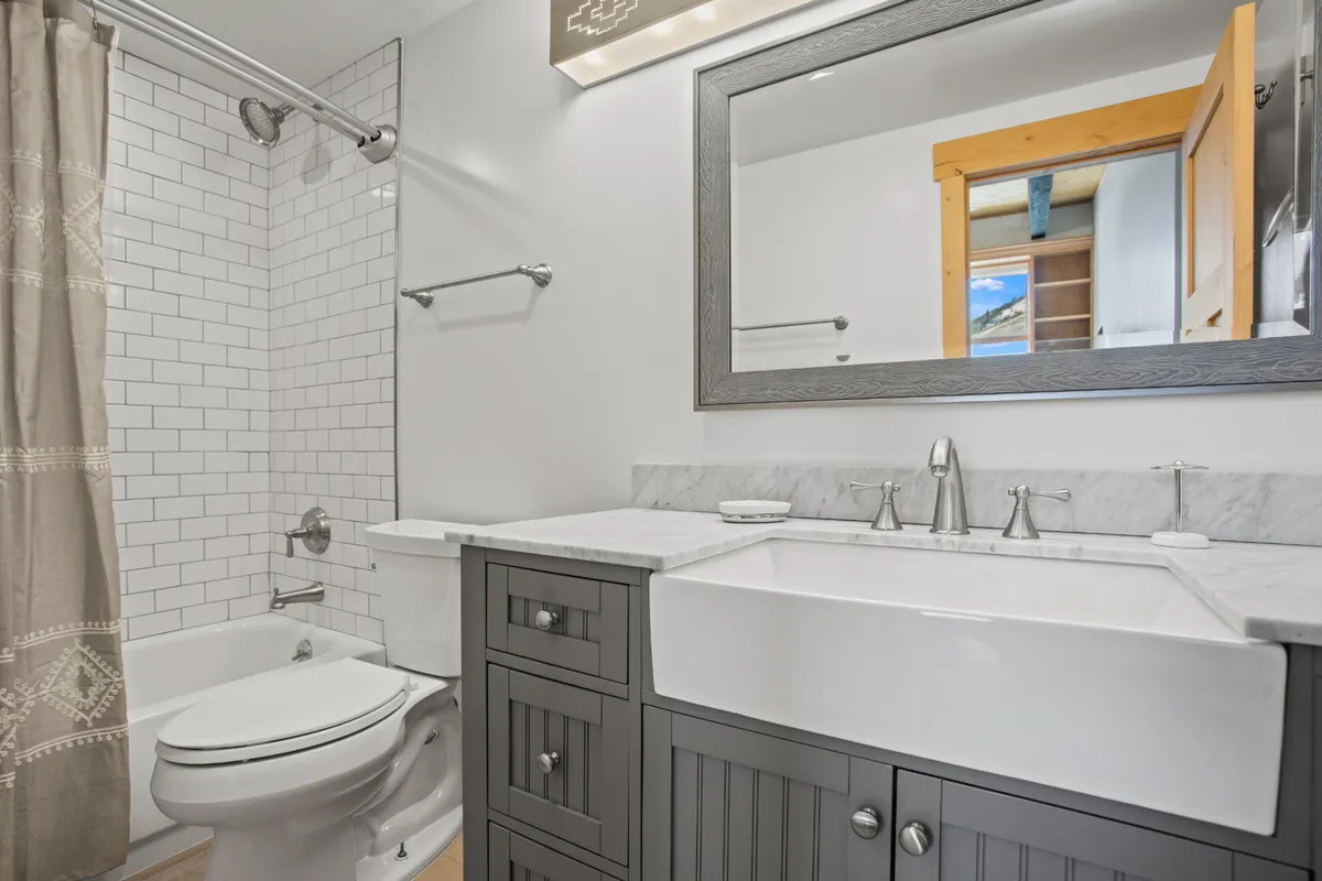 This image features a modern, spa-like bathroom with a dual sink vanity, large mirror, and a spacious shower with tile surround. Natural light filters in through a wooden door window, and the inviting setup suggests a focus on relaxation, refreshment, and self-care.