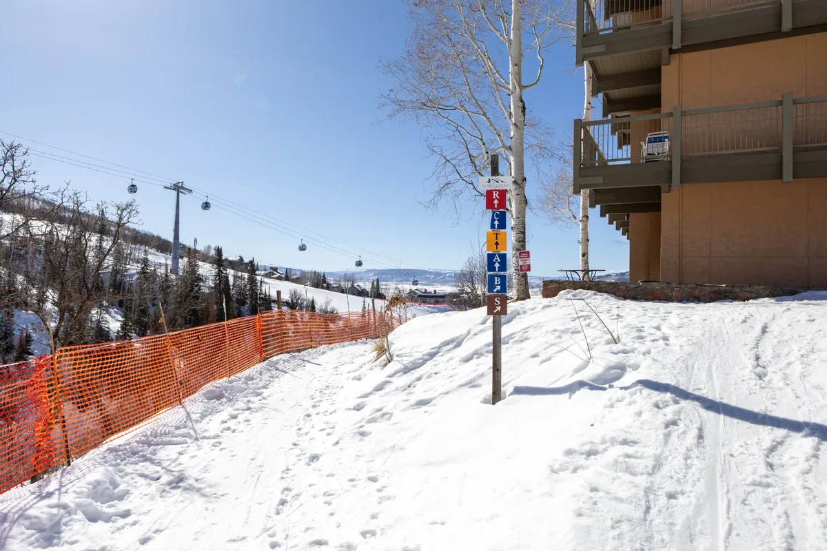 A snow-covered pathway leads directly to a well-marked ski trail, with a mountain view in the background and part of the building complex visible to the side. The scene is lively with fresh snow, ski trail signs, and an inviting atmosphere that highlights both ski access and the property's proximity to the slopes.