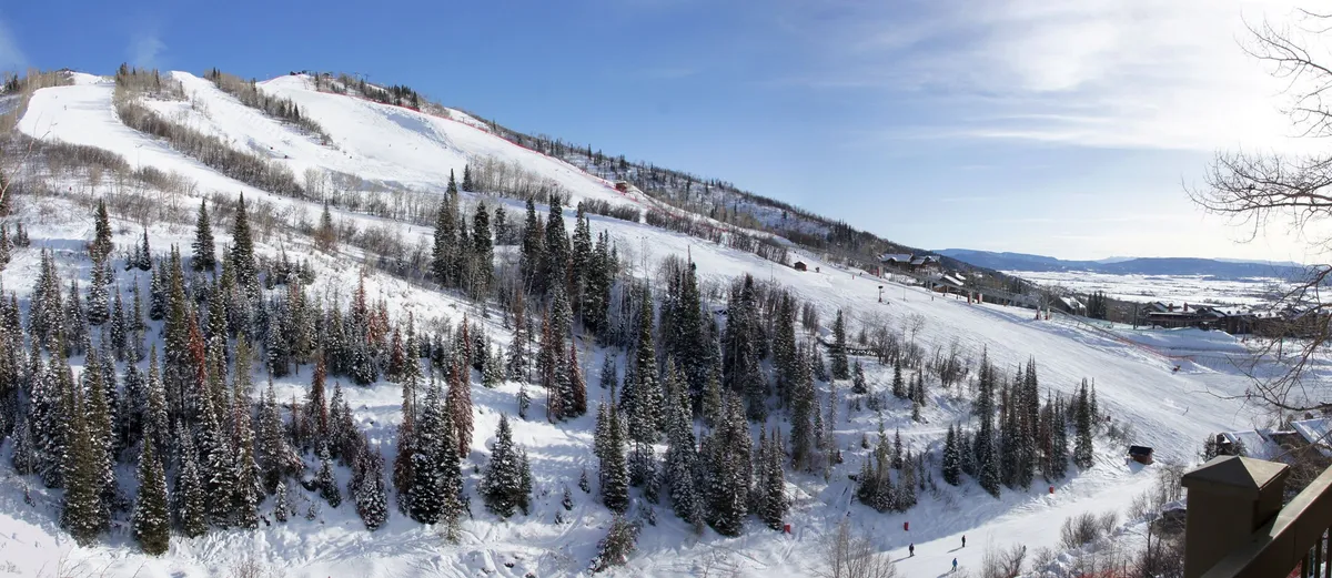 The image captures a snowy mountain landscape with skiers traversing the slopes, surrounded by evergreen trees and distant mountain peaks. The lively winter scene is invigorating, with clear skies and visible resort amenities in the distance.