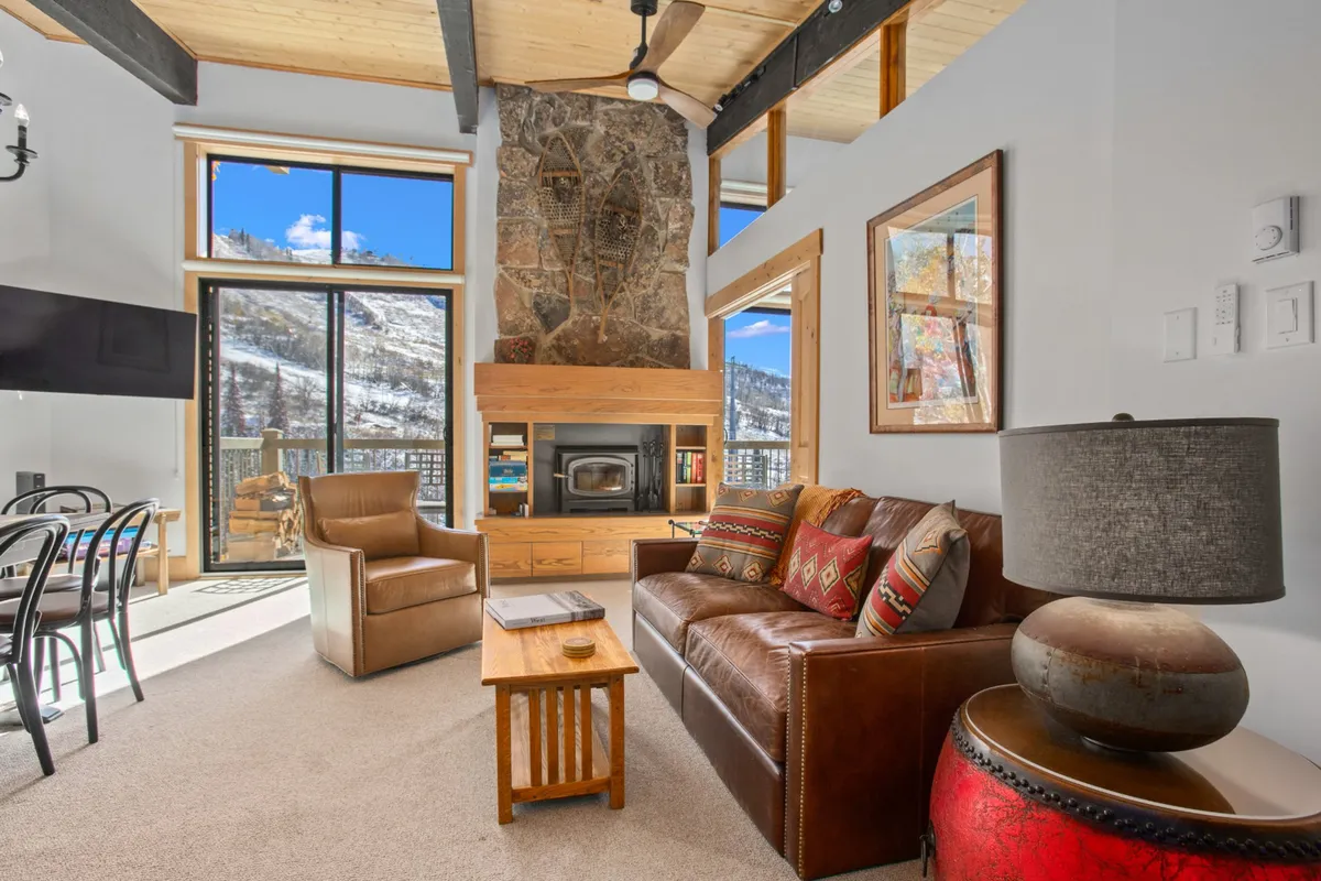 A sunlit living room featuring a stone fireplace as a focal point, with a comfortable sofa positioned to take in the scenery through expansive windows that offer breathtaking mountain views. The warm textures of wood and stone, paired with plush furnishings, create a cozy, inviting space ideal for relaxing and social gatherings.