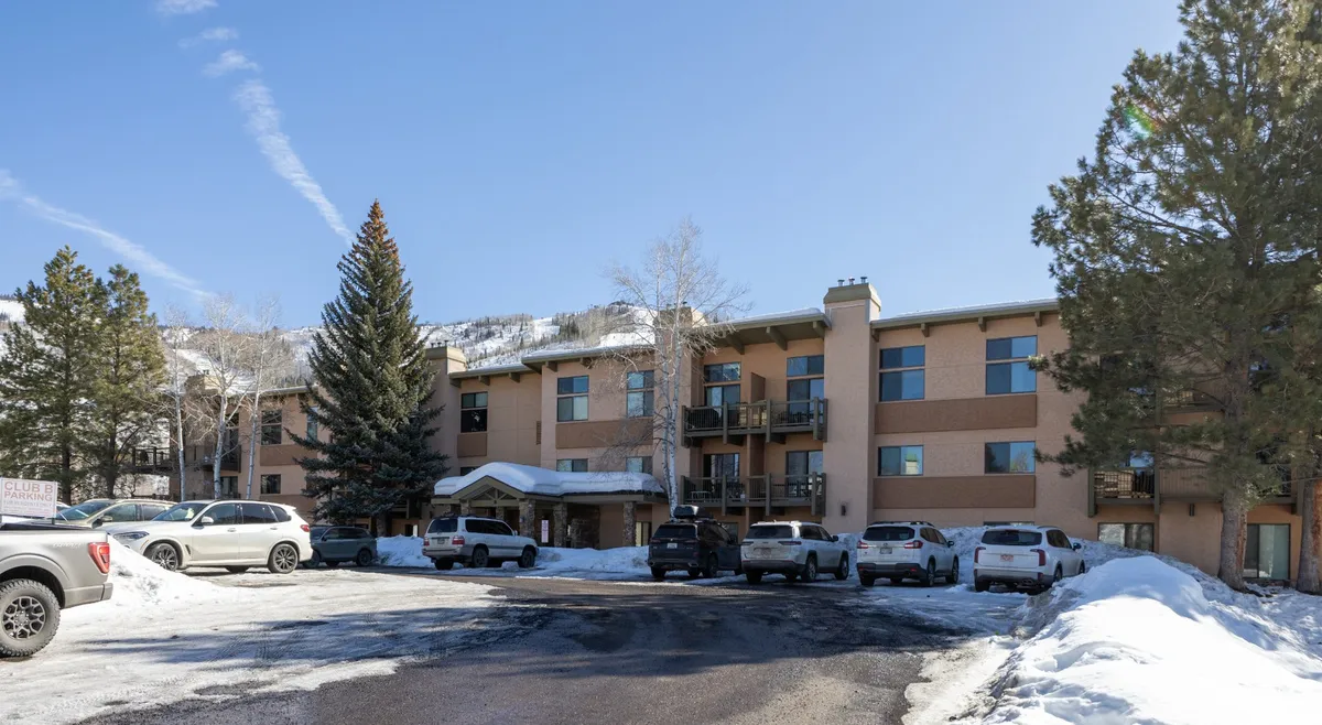 The image features a snow-covered exterior with pine trees, balconies, and communal outdoor areas. The setting feels tranquil and inviting, with hints of amenities and gathering spaces for groups to enjoy outdoor relaxation or community events.