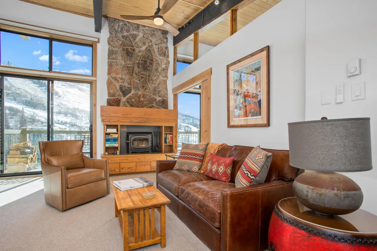 The image showcases a beautifully remodeled condo living room on the penthouse level. A glowing fireplace is the centerpiece, flanked by a cozy sofa with inviting decorative cushions and a rustic wooden coffee table. Large windows reveal glimpses of mountain vistas and capture the warm glow of the sunset, enveloping the space in natural light and creating a tranquil atmosphere ideal for relaxation after a day on the slopes.