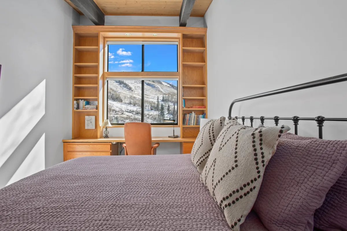 A serene bedroom with a cozy bed, textured throw pillows, a wooden desk and chair positioned by a large window, and sweeping mountain views. The overall ambiance is warm and restful, ideal for relaxation or work, echoing the retreat theme.