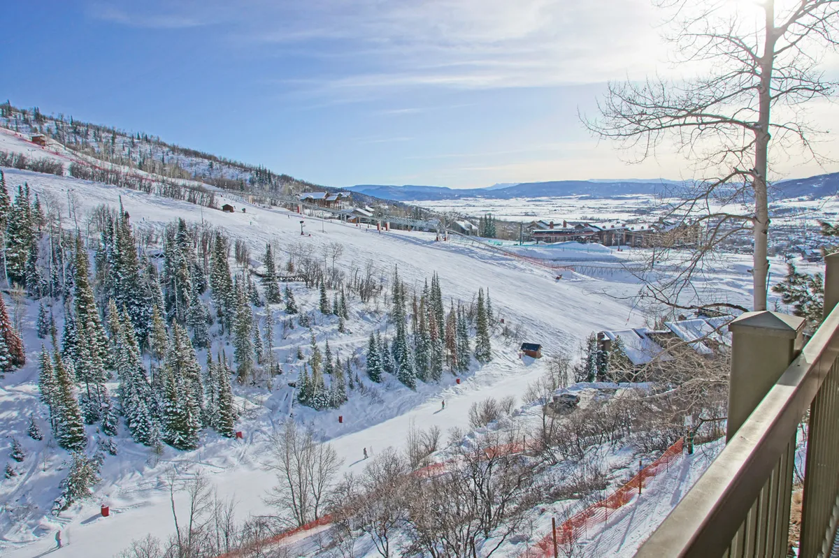 A lively winter scene featuring snow-covered slopes, pine trees, several skiers in motion, and distant mountain ranges under a clear blue sky. The vantage point suggests an elevated position, possibly from a balcony or upper floor, capturing the excitement and panoramic beauty of Steamboat's ski area.