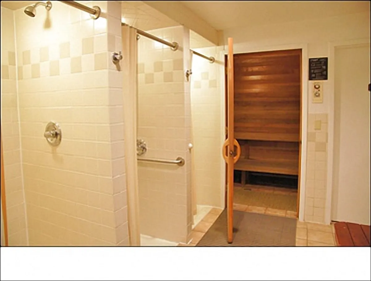A calming interior view of a spa area featuring tiled walls, individual shower stalls, and the entrance to a warm, inviting sauna. The space exudes a restorative spa-like ambiance, ideal for relaxation and self-care.