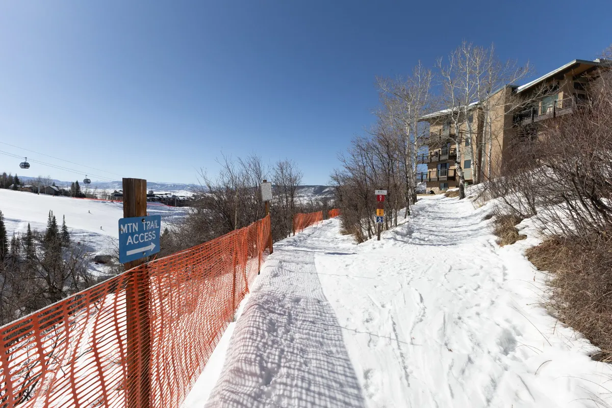 This exterior image showcases a snow-covered path leading directly to the building, with a mountain trail access sign visible and the structure in the background. The surrounding trees and fresh snow highlight the seamless connection between the property and the ski trails.
